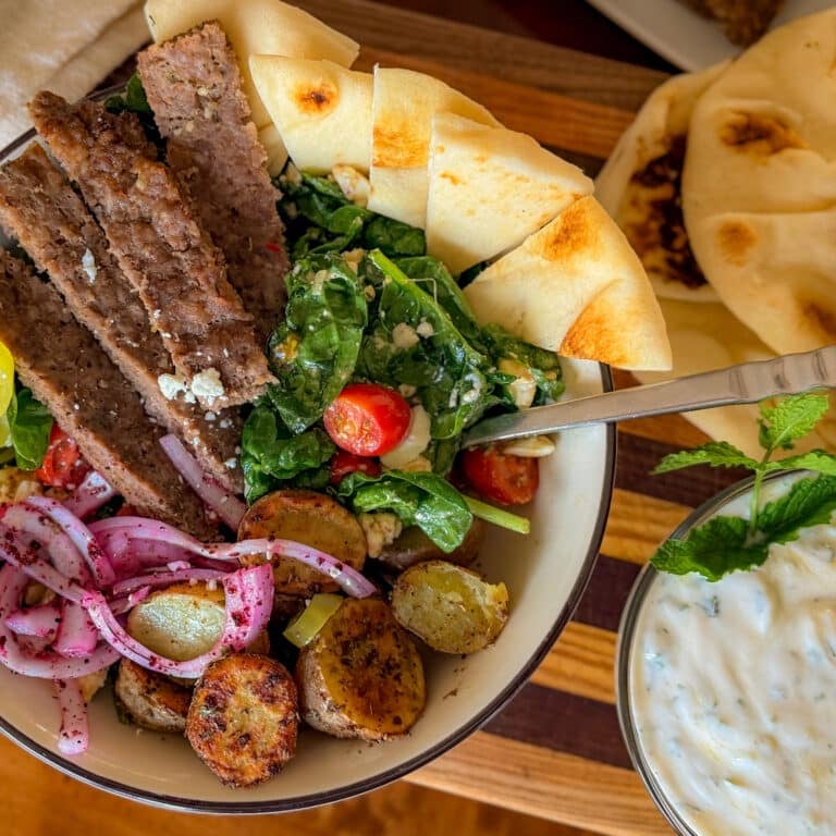 Close Up of a Homemade Gyro Bowl Filled with Seasoned Beef and Chicken Gyro Meat, Golden Roasted Potatoes, Fresh Spinach, Tomatoes, Feta and Sumac Red Onions, with Tzatziki on the Side.