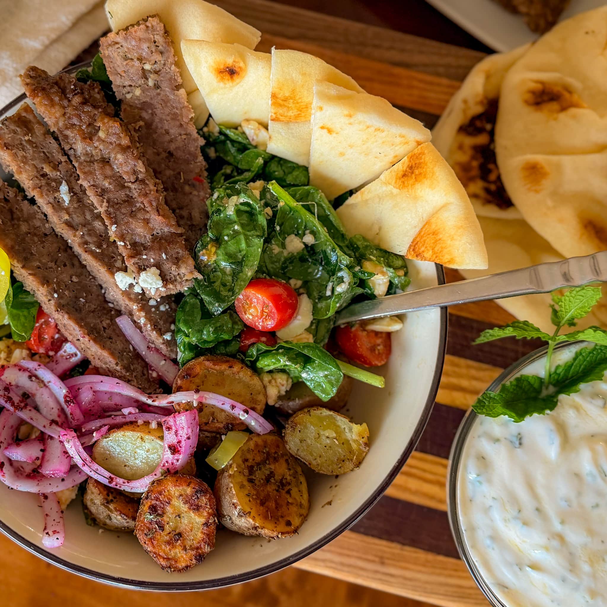 Gyro Bowls with Roasted Potatoes and Sumac Onions Close Up of a Homemade Gyro Bowl Filled with Seasoned Beef and Chicken Gyro Meat, Golden Roasted Potatoes, Fresh Spinach, Tomatoes, Feta and Sumac Red Onions, with Tzatziki on the Side.